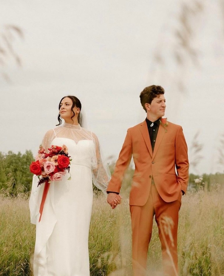 Bride and groom standing in an outdoor field, with the groom wearing a custom burnt orange suit and the bride in a white wedding dress, holding a bouquet of red and pink flowers.