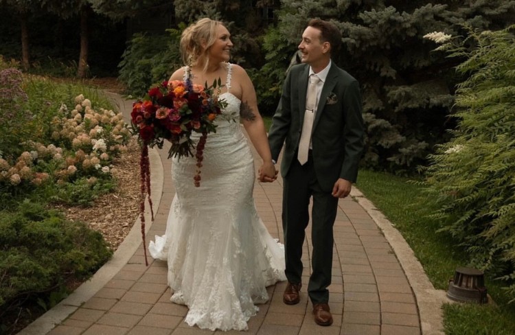 Bride and groom walking through a garden, with the groom wearing a custom dark green suit and the bride in a white lace wedding dress, holding a vibrant bouquet of red and orange flowers.