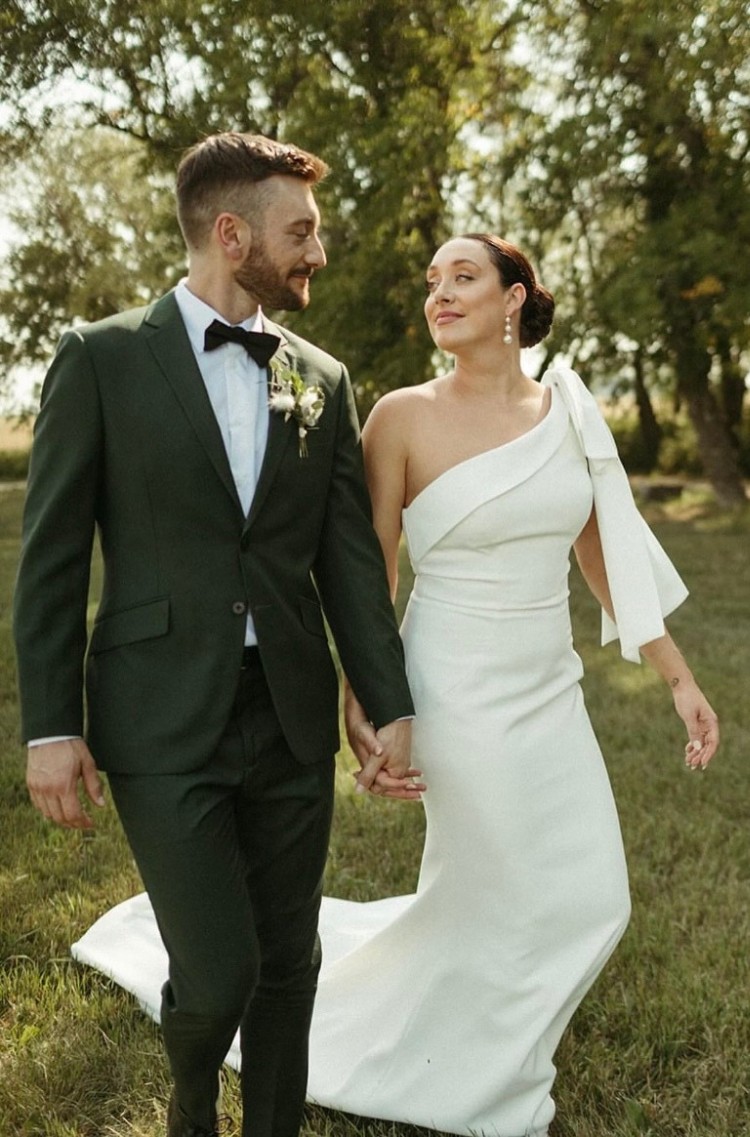 Bride and groom walking hand in hand in a grassy outdoor setting, with the groom wearing a custom dark green suit and the bride in a white one-shoulder wedding dress.