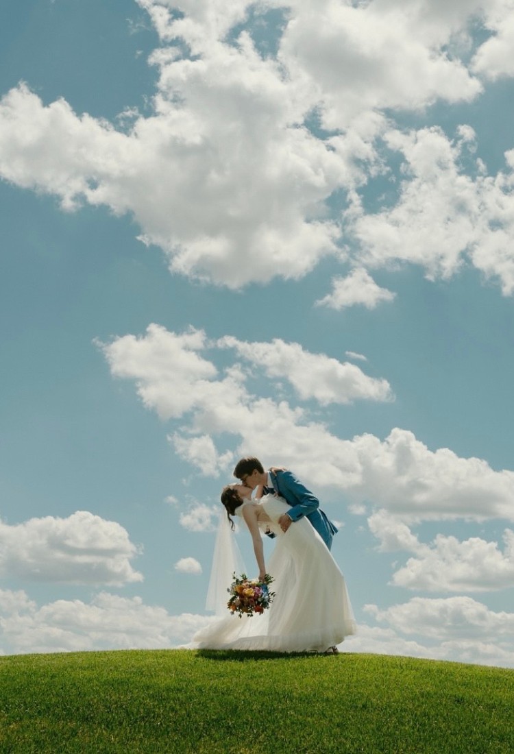 Bride and groom sharing a kiss on a grassy hill under a bright blue sky with clouds, with the groom wearing a custom blue suit and the bride in a white wedding dress holding a bouquet of colorful flowers.