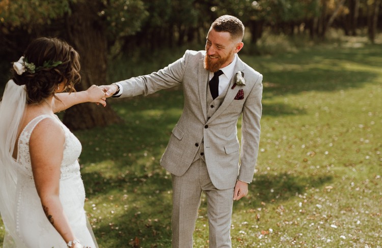 Groom wearing a light grey custom suit holding hands with the bride in a sunny outdoor setting, smiling and sharing a moment.