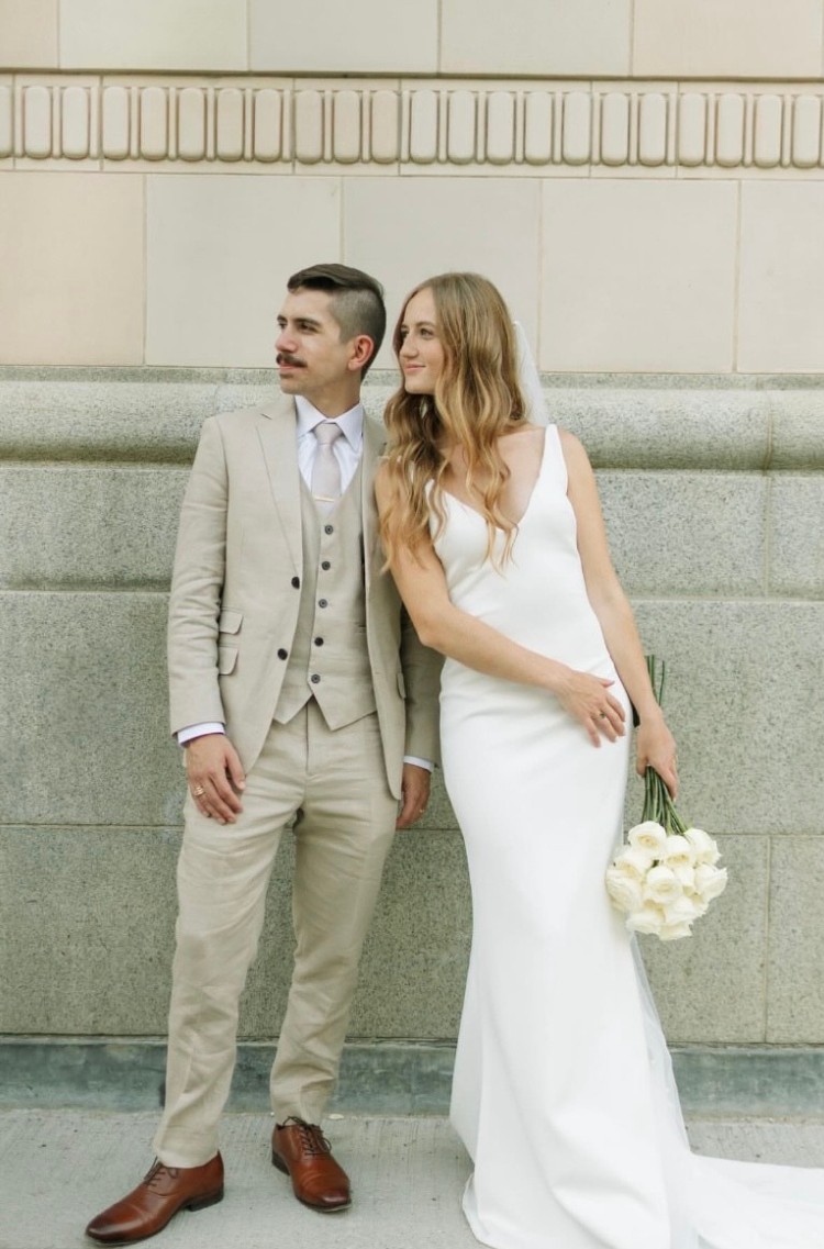 Groom in a light beige three-piece custom suit standing next to the bride in a sleek white wedding dress holding a bouquet of white roses.