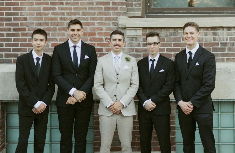 Groom in a beige custom suit with groomsmen wearing classic black suits, posing against a brick wall.