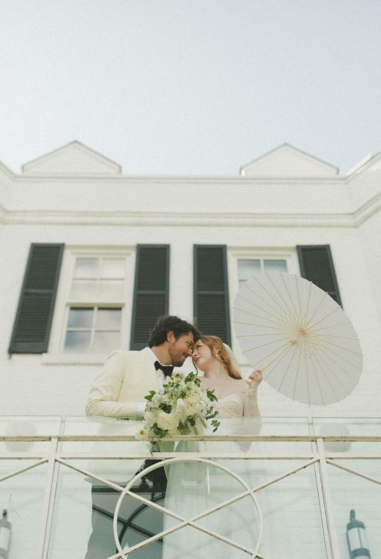 Groom in a white tuxedo and bride with a parasol, sharing a romantic moment on a balcony.