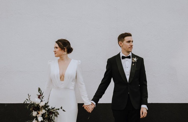 Bride in a white gown holding hands with the groom in a classic black tuxedo, standing against a black and white background.