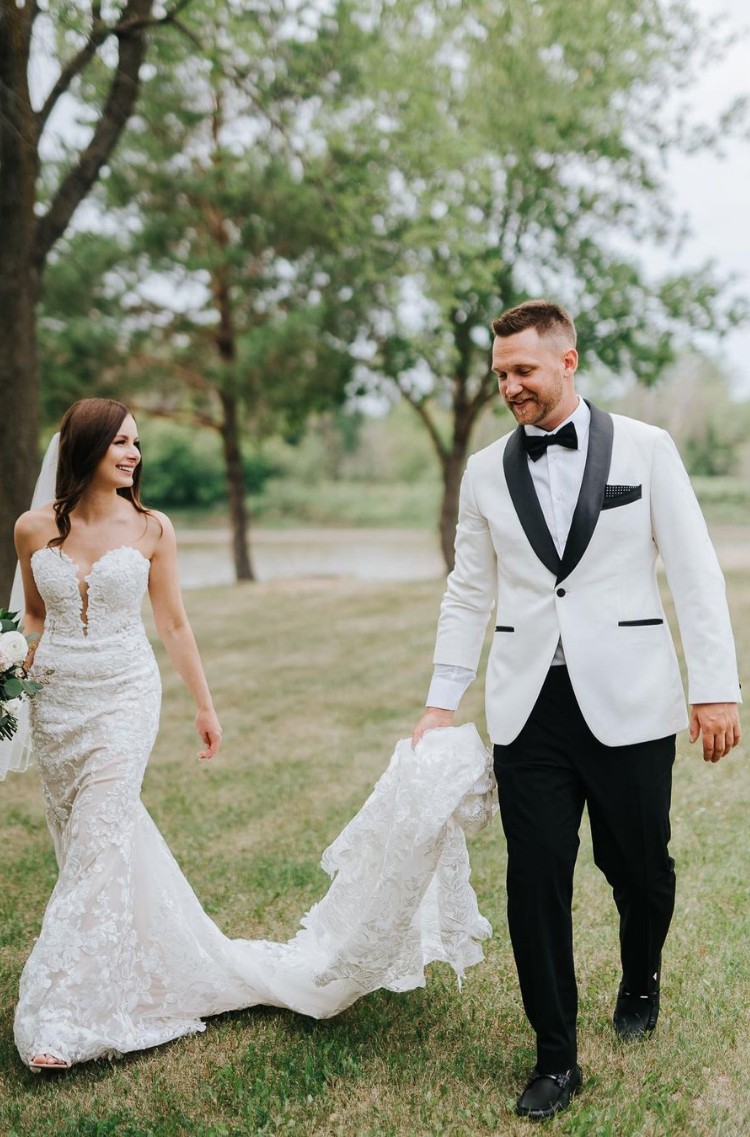 Groom in a white tuxedo jacket with black accents holding the bride's lace train as they walk together outdoors