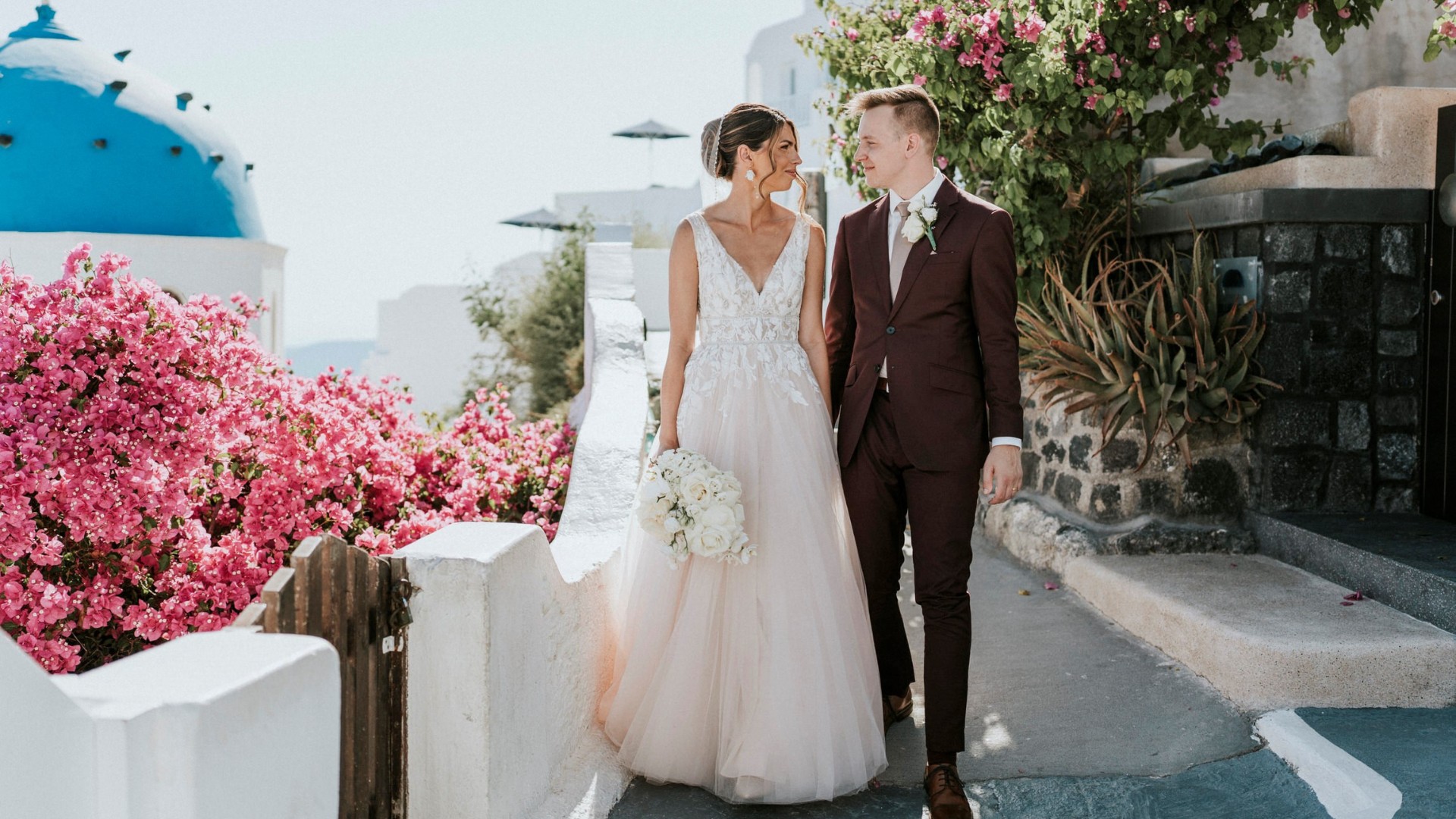 Bride and groom walking together in a picturesque Greek setting with bright pink flowers and a blue-domed building.