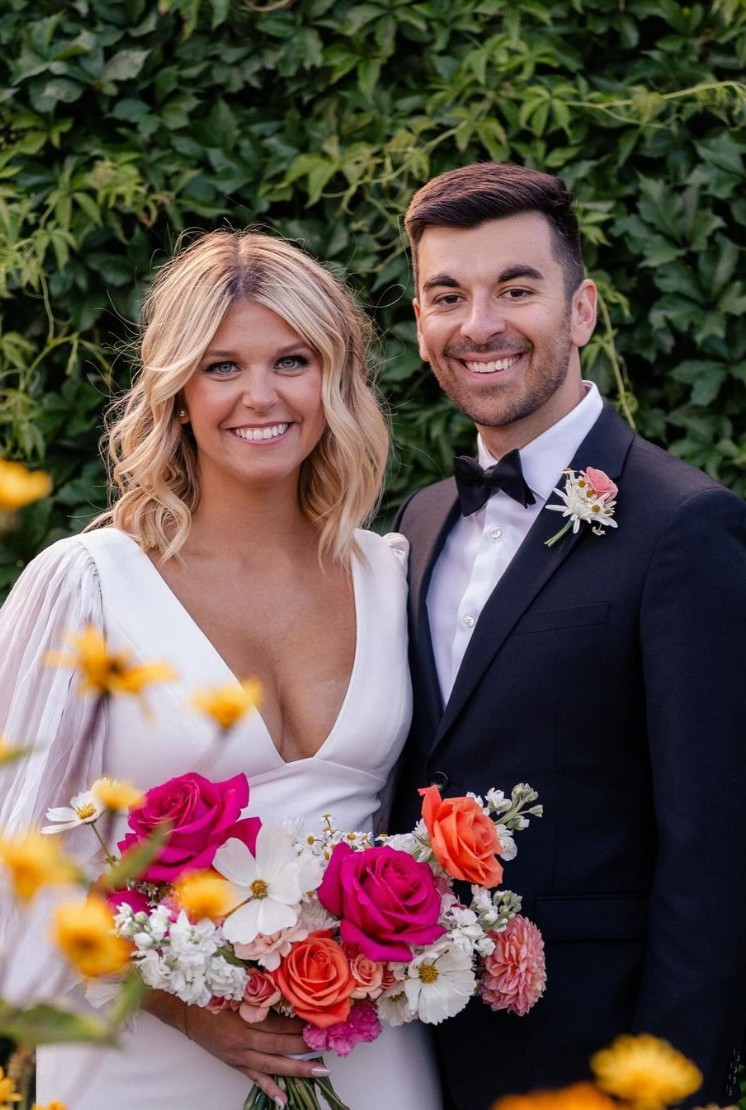 Bride and groom smiling, with the groom wearing a black tuxedo and the bride holding a vibrant floral bouquet.