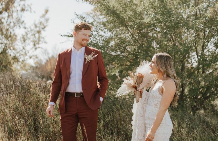 Groom in a burnt orange suit from Eph Apparel, standing with his bride in a natural outdoor setting.