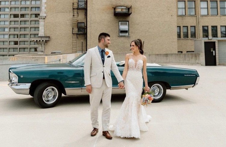 Bride and groom standing in front of a classic blue car, with the groom in a beige suit and the bride in a lace wedding dress.