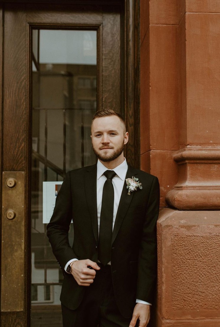 Groom in a classic black suit standing by a vintage-style door, wearing a boutonniere.