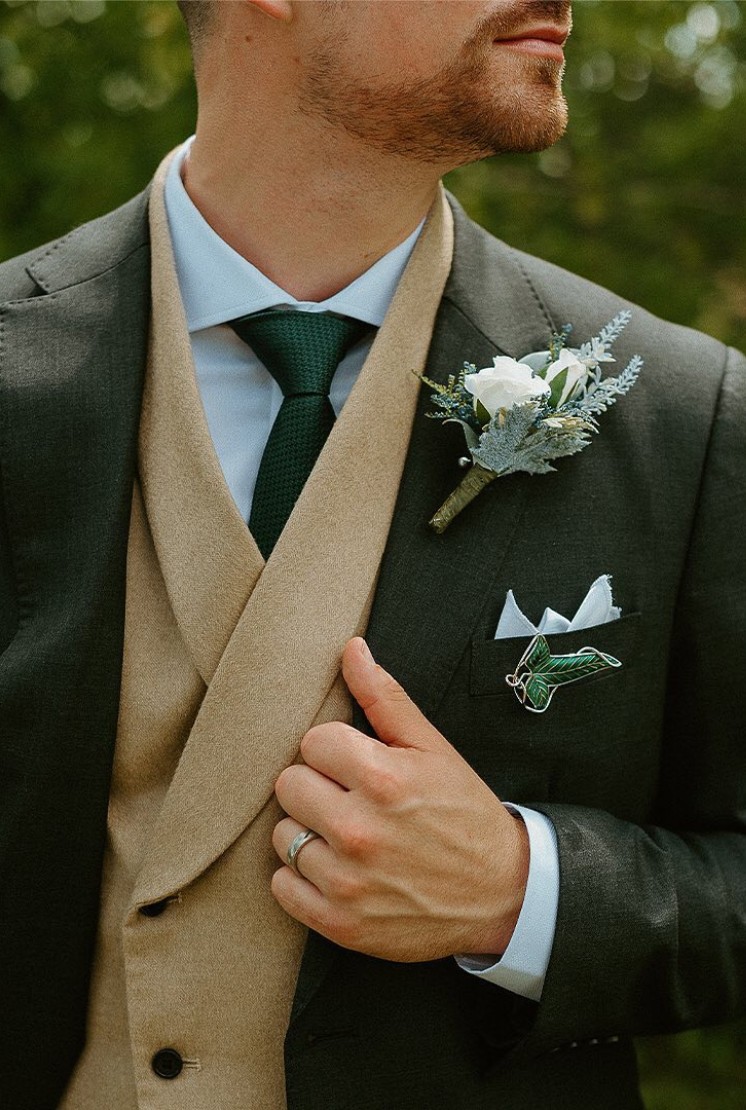 Close-up of a groom wearing a dark green suit with a beige shawl lapel vest and matching green tie, featuring a boutonniere and pocket square.