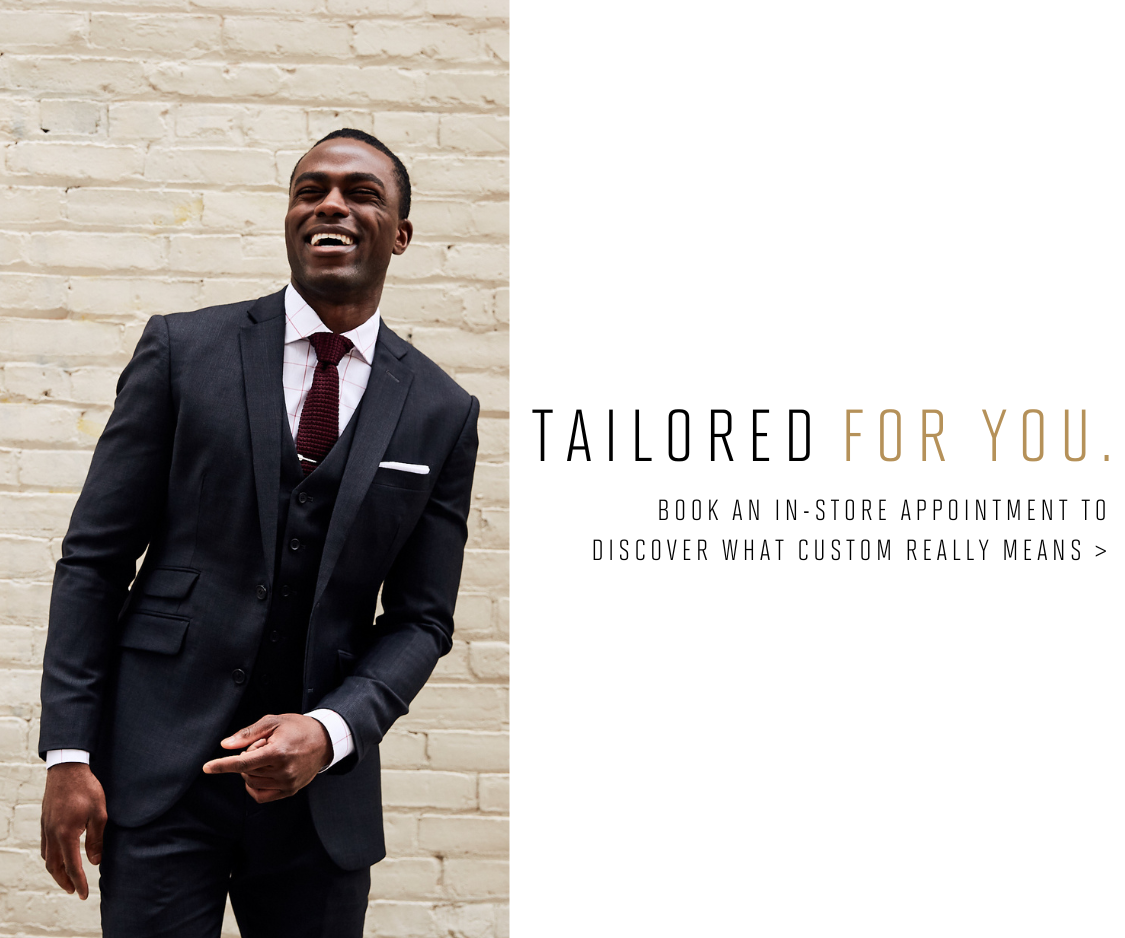 Man in a custom black suit smiling against a brick wall with the text 'Tailored for You' and call to action to book an in-store appointment for a custom experience.