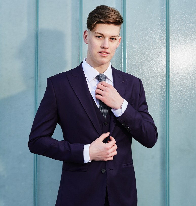 A young man in a custom purple suit, white dress shirt, and gray tie, styled by Eph Apparel, stands confidently in front of a teal background, showcasing a polished look for Manitoba graduates.