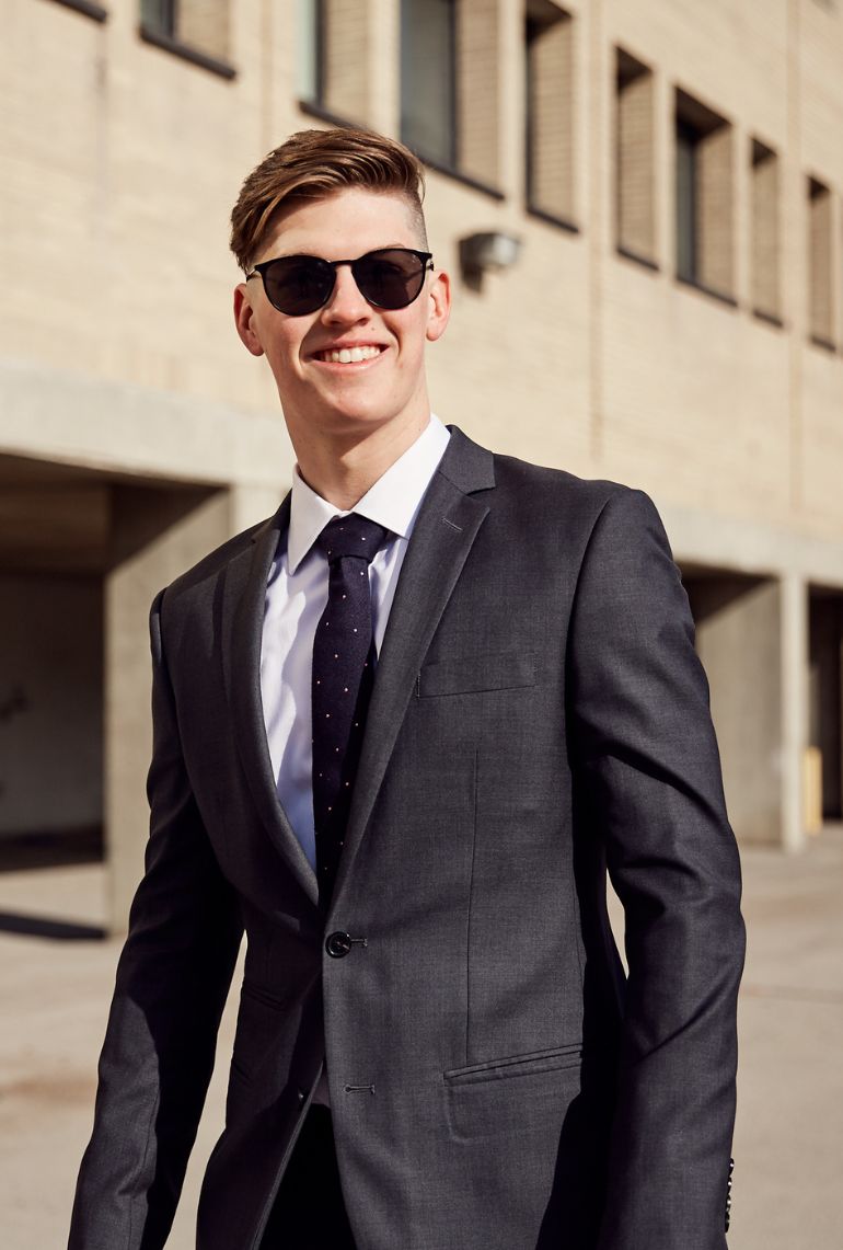 A young man in a custom purple suit, white dress shirt, and gray tie, styled by Eph Apparel, stands confidently in front of a teal background, showcasing a polished look for Manitoba graduates.